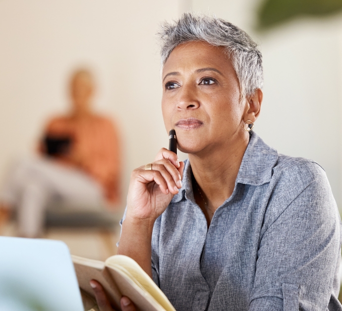 Woman with gray hair sitting at desk holding notebook and looking pensive